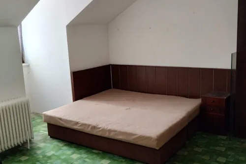 Původní vybavení podkrovního pokoje dřívějšího ubytování na Hlinkách 28a: postel u tmavého obložení a zelený vzorovaný koberec, srpen 2025