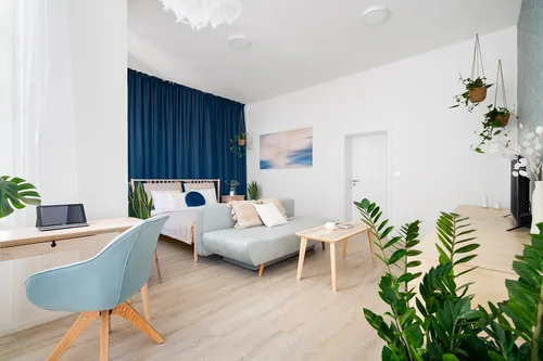 Apartmán s postelí před modrým závěsem, pohovkou a pracovním stolem, červenec 2023