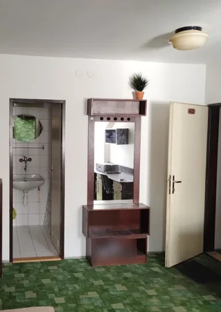 Pokoj dřívějšího ubytování na Hlinkách 28a před převzetím domu: zelený vzorovaný koberec, tmavý nábytek, umyvadlo v malé koupelně a dveře s číslem pokoje, 2025